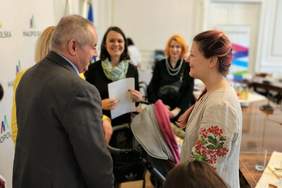 Pokaż zdjęcie: Na pierwszym planie niewidomy mężczyzna wita się z młodą kobietą. W tle uśmiechnięte kobiety.