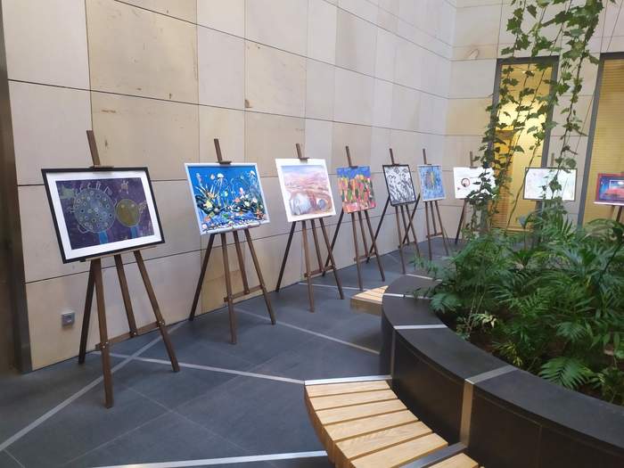 Pokaż zdjęcie: Na tle kamiennej ściany stoją sztalugi, a na nich są wyeksponowane obrazy, po prawe stronie zdjęcia kompozycja z roślin zielonych