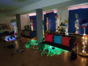 Pokaż zdjęcie: wnętrze dużej sali, w półmroku widać stolik z włączonym projektorem dwie czarne ławki z kolorowymi poduszkami, na podłodze leży oświetlenie ledowe zmieniające kolor w sali rozstawionych jest 7 kukieł z maseczkami i kolorowymi nakryciami głow