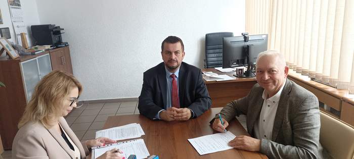 Pokaż zdjęcie: Przy stoliku dwaj mężczyźni i kobieta - przedstawiciele Powiatu Wschowskiego i PFRON, którzy podpisują umowę, są uśmiechnięci i ubrani na galowo