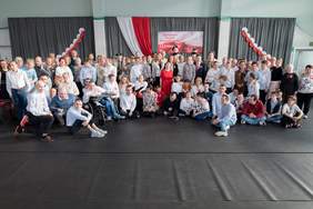 Pokaż zdjęcie: Duża grupa mieszkańców Domu Pomocy Społecznej w Białymstoku oraz zaproszonych gości pozuje wspólnie do zdjęcia podczas obchodów Narodowego Święta Niepodległości. W tle widać biało-czerwone dekoracje oraz baner z napisem „Narodowe Święto Niepodległości – 11 Listopada”.