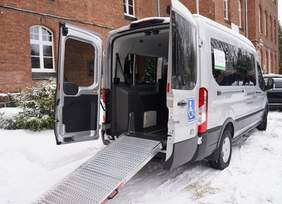 Pokaż zdjęcie: Specjalistyczny bus przystosowany do przewozu osób z niepełnosprawnościami, zaparkowany przy ceglanym budynku. Tylne drzwi są otwarte, wysunięta metalowa rampa umożliwia wjazd wózka. Na pojeździe oznaczenie dostępności.