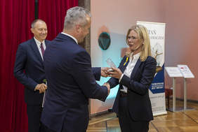 Pokaż zdjęcie: Mężczyzna w granatowym garniturze białe koszuli, wręcza kobiecie statuetkę. Kobieta w blond włosach ubrana  w granatowy damski biały garnitur, białą koszulę, okulary, uśmiecha się przejmując nagrodę.