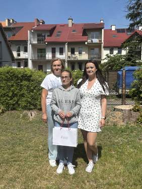 Pokaż zdjęcie: Trzy osoby stoją na trawniku przed budynkiem mieszkalnym z czerwonym dachem. Osoba znajdująca się pośrodku trzyma torbę z upominkiem. Fotografia została wykonana w słoneczny dzień, a w tle widoczne są krzewy, ogrodzenie oraz wielorodzinny budynek mieszkalny. Uczestnicy pozują do pamiątkowego zdjęcia na świeżym powietrzu.