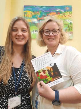 Pokaż zdjęcie: Stoją dwie panie i uśmiechają się szeroko. Jedna z pań trzyma w dłoni ulotkę informacyjną. Na ścianie za nimi wisi kolorowy obrazek. 