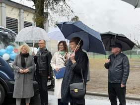 Pokaż zdjęcie: Pięć osób stoi na zewnątrz w deszczowy dzień przed niebieskim busem udekorowanym biało-niebieskimi balonami. Po lewej kobieta w szarym płaszczu i czarnym szaliku. Obok niej mężczyzna w okularach i ciemnej kurtce z jasnym parasolem we wzory. W centrum kobieta w kremowym płaszczu i zielonym szaliku trzyma niebieską teczkę. Na prawo kobieta w ciemnym płaszczu z granatowym parasolem i torebką, a obok niej mężczyzna w czapce i skórzanej kurtce pod czarnym parasolem.