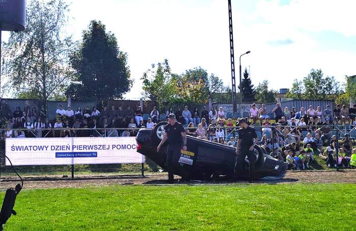 Na boisku sportowym, na bieżni leży, wywrócony kołami do góry, samochód osobowy - upozorowano wypadek.. Obok niego stoją dwaj policjanci. Po prawej stronie zdjęcia, na płocie, wisi baner z napisem "Światowy Dzień Pierwszej Pomocy" Zakład Karny w Potulicach. W tle, za płotem, siedzi duża grupa widzów.