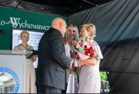 Pokaż zdjęcie: Trzy osoby stoją blisko siebie, dwie z nich wręczają kwiaty i grawerton kobiecie w jasnej sukience.