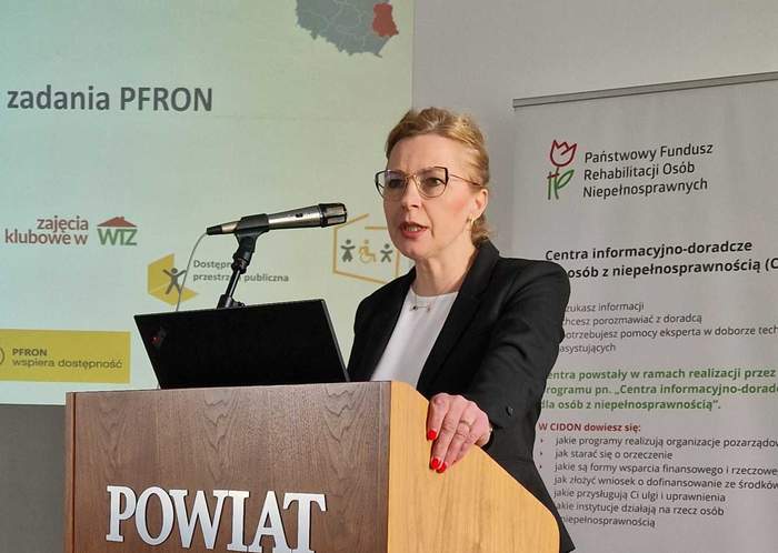 Pokaż zdjęcie: Przy mównicy stoi kobieta blondynka z upiętymi włosami w okularach, lewą rękę trzyma na mównicy.