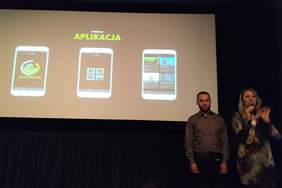 Pokaż zdjęcie: Sala w Kinie pod Baranami. Na ekranie wyświetlają się informacje dotyczące aplikacji AudioMovie. Przed ekranem stoją Piotr Franiek i Regina Mynarska, którzy zapraszają na seans.