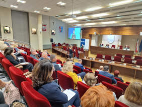Pokaż zdjęcie: Widok na sale konferencyjną z boku i z góry, widać uczestników, za stołem prezydialnym na przodzie siedzą 4 osoby