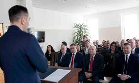 Pokaż zdjęcie: Zdjęcie sali, w której na krzesełkach siedzą zaproszeni goście, elegancko ubrani. Przy mównicy stoi mężczyzna tyłem do zdjęcia, przemawia do gości.