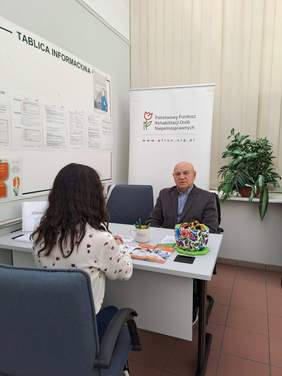 Pokaż zdjęcie: Przy stoliku na którym leżą ulotki i katalogi, siedzą kobieta i mężczyzna. W tle baner z logo PFRON.