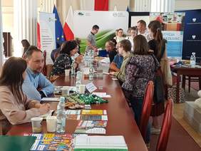 Duża sala, na niej targi pracy, część osób siedzi za stołami, pozostali chodzą