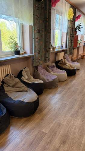 Pokaż zdjęcie: Jasne pomieszczenie z dużymi oknami i pufami do siedzenia ustawionymi wzdłuż ściany. Wnętrze pełni funkcję strefy relaksu dla uczestników warsztatu.