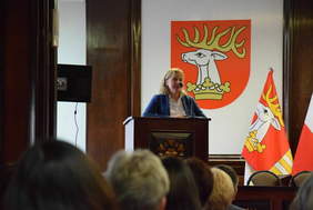 Pokaż zdjęcie: Zdjęcie mównicy przy której stoi kobieta blondynka w niebieskim garniturze.