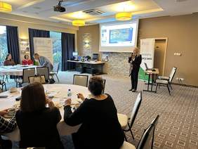 Pokaż zdjęcie: Sala konferencyjna. Po lewej stronie Stoi baner CIDON obok wisi ekran do rzutnika z prezentacją. Pod nim stolik z urządzeniami elektronicznymi. Po prawej stronie stoi kobieta w blond włosach ubrana na ciemno z apaszką. Trzyma w ręku mikrofon i mówi 