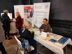 Pokaż zdjęcie: Przy stoisku Łódzkiego Oddziału PFRON siedzą naprzeciwko siebie dwie kobiety. Patrzą na siebie i rozmawiają ze sobą. Na stole leżą różne materiały reklamowe, informatory, broszury, ulotki. 