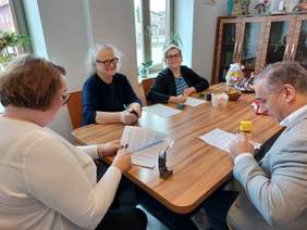 Pokaż zdjęcie: Przy stole sidzi mężczyzna trzyma długopis i czyta umowę. Naprzeciw siedzą dwie uśmiechnięte kobiety w okularach, jedna z nich podpisuje umowę druga trzyma pieczątkę. Obok kobieta w krótszych włosach i w białej bluzce podpisuje dokument.