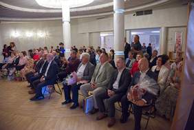 Pokaż zdjęcie: Publiczność siedząca na sali podczas uroczystości; osoby w eleganckich strojach, część trzyma kwiaty i upominki, widoczne kolumny, żyrandol i jasne oświetlenie wnętrza.