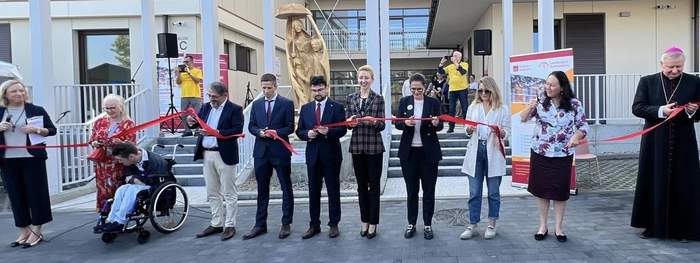 Pokaż zdjęcie: Przed nowoczesnym budynkiem stoją elegancko ubrane osoby trzymające w rękach wstęgę i przecinające ją. Wśród nich ksiądz i osoba na wózku. Za nimi drewniany pomnik kobiety z dzieckiem.