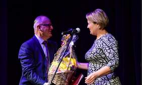 Pokaż zdjęcie: Na scenie stoi mężczyzna w okularach oraz kobieta, kobieta wręcza mężczyźnie duży kosz cukierków. Kosz jest przyozdobiony kokardą