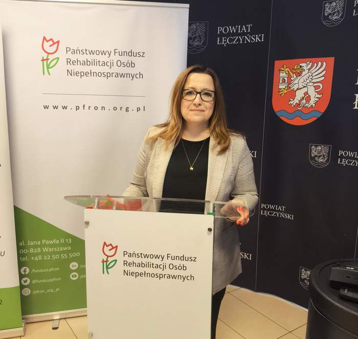 Pokaż zdjęcie: Przy mównicy stoi kobieta z ciemnymi półdługimi włosami w okularach.