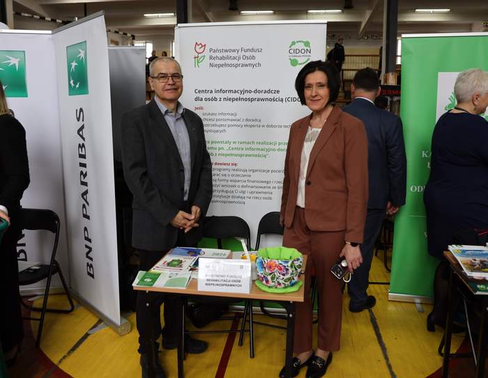 Pokaż zdjęcie: Przy stoliku na którym leżą ulotki, stoją dwie osoby, w tle rollup z logo PFRON.