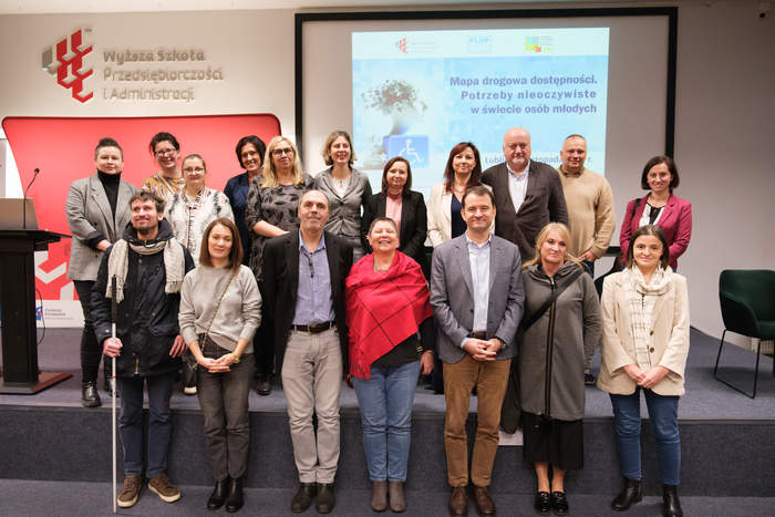 Pokaż zdjęcie: Na scenie w dwóch rzędach stoi duża grupa osób. Uśmiechają się do pamiątkowego zdjęcia.