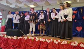 Pokaż zdjęcie: Grupa uczestników stoi w rzędzie na scenie przy mikrofonach, prezentując część artystyczną. W tle widoczna jest dekoracja z czerwonymi sercami i granatowy baner okolicznościowy.
