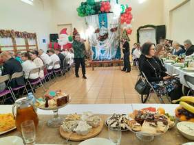 Pokaż zdjęcie: W dużej sali stoją stoły, które są ustawione w podkowę. Przy stołach siedzą ludzie, którzy wspólnie biesiadują. Dwie osoby stoją i robią pamiątkowe zdjęcia. 