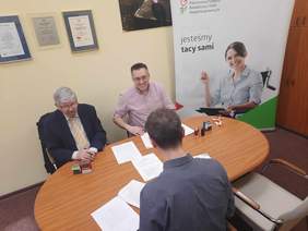 Pokaż zdjęcie: W gabinecie znajduje się stół, przy którym siedzi trzech panów. Zwierają umowę o dofinansowanie. W pokoju jest duży baner z napisem: Wszyscy jesteśmy tacy sami. 