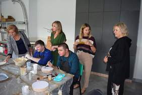 Pokaż zdjęcie: W pracowni przy stole dwóch młodych mężczyzn dekorują ceramikę, cztery kobiety stoją i podziwiają ich prace