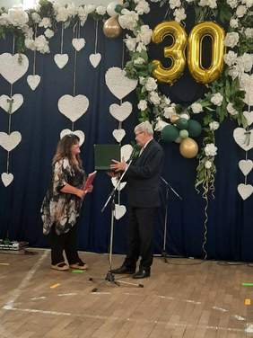 Pokaż zdjęcie: Na środku sali mężczyzna wręcza kobiecie grawer. Pani uśmiecha się szeroko. Na ścianie ze złotych balonów zrobiona jest liczba "30". 