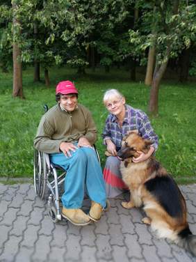 Pokaż zdjęcie: Od lewej mężczyzna na wózku, obok niego kobieta, jego matka, szefowa stowarzyszenia „krok dalej” oraz ich pies, owczarek niemiecki. Zdjęcie zrobione na polanie. 