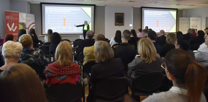 Pokaż zdjęcie: Około trzydzieści osób siedzi w dużej sali. Z przodu stoi kobieta wskazująca jedną ręką wykres wyświetlany na dużym ekranie. W sali są dwa ekrany, a po obu ich stronach ustawione zostały banery PFRON oraz Muzeum Pierwszych Piastów na Lednicy.