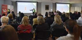 Pokaż zdjęcie: Około trzydzieści osób siedzi w dużej sali. Z przodu stoi kobieta wskazująca jedną ręką wykres wyświetlany na dużym ekranie. W sali są dwa ekrany, a po obu ich stronach ustawione zostały banery PFRON oraz Muzeum Pierwszych Piastów na Lednicy.