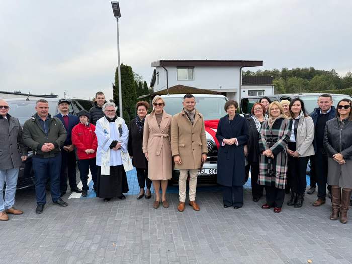 Pokaż zdjęcie: Grupa kilkunastu osób, wśród których jest również ksiądz, stoi na tle trzech mikrobusów. Z tyłu znajdują się domy jednorodzinne oraz fragment lasu. 