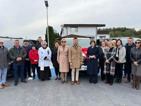 Pokaż zdjęcie: Grupa kilkunastu osób, wśród których jest również ksiądz, stoi na tle trzech mikrobusów. Z tyłu znajdują się domy jednorodzinne oraz fragment lasu. 