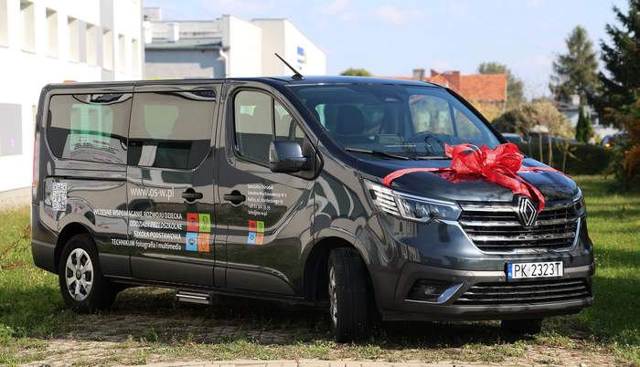 Pokaż zdjęcie: Czarny bus udekorowany czerwoną kokardą na masce oznaczony jest nazwą i adresem SOSW nr 2 w Kaliszu, jak również informacją o placówkach wchodzących w skład Ośrodka. W tle duży biały budynek oraz zabudowania jednorodzinne.