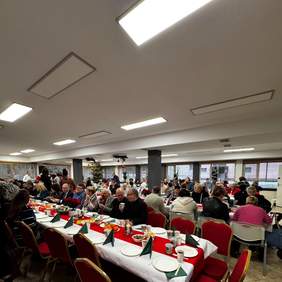 Pokaż zdjęcie: Przy długich stołach siedzą ludzie. Rozmawiają ze sobą, jedzą i piją. Stoły są zastawione świątecznymi potrawami. 