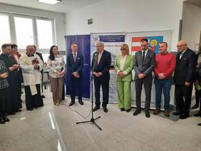 Pokaż zdjęcie: W półkolu stoją ludzie. Na środku ustawiony jest mikrofon. Przemawia do niego dyrektor Łódzkiego Oddziału PFRON. Wszyscy są elegancko ubrani. 