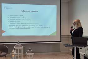 Pokaż zdjęcie: Na zdjęciu ekran z prezentacją dotyczącą Systemu Obsługi Dofinansowań dla pracodawców - omawiane Schorzenia specjalne przy orzeczeniach o niepełnosprawności. Po prasowej stronie pracownica Oddziału Zachodniopomorskiego PFRON w jasnych włosach, w ciemnej garsonce z mikrofonem w ręku.