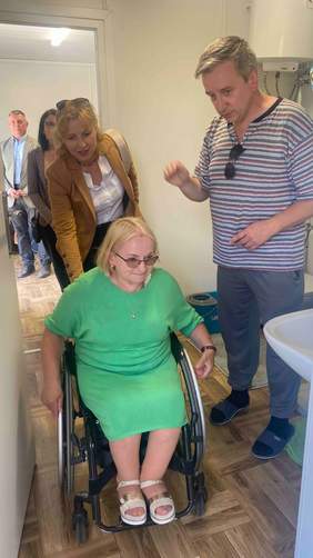 Pokaż zdjęcie: Na pierwszym planie kobieta na wózku inwalidzkim, z tyłu stoi kobieta, która pomaga, aby przejechał wózek, po prawej stronie stoi mężczyzna. Zdjęcie w kontenerze mieszkalnym 