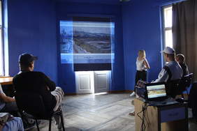 Pokaż zdjęcie: Uczestnicy ŚDS Caritas podczas zajęć w "Szafirowej pracowni" - dorosłe osoby z niepełnosprawnościami oglądają film wyświetlony na ekranie, w tle niebieska ściana