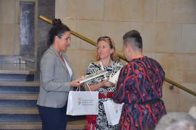 Pokaż zdjęcie: 3 zwrócone ku sobie kobiety trzymające w dłoniach torebki prezentowe PFRON i oprawione w ramki dyplomy. W tle marmurowe schody.