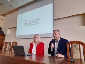 Pokaż zdjęcie: Za stołem konferencyjnym siedzi blondynka w czerwonej marynarce oraz mężczyzna w marynarce z mikrofonem w ręku. Na ścianie slajd z logo PFRON.