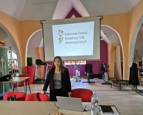 Pokaż zdjęcie: Prelekcja, wystąpienie kobiety z długimi włosami, w tle slajd z prezentacją i logo Funduszu.
