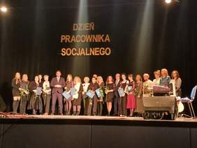 Pokaż zdjęcie: Na scenie Grajewskiego Centrum Kultury stoi grupa kobiet, która została wyróżniona i nagrodzona z okazji obchodów Dnia Pracownika Socjalnego. Wszystkie trzymają w rękach kwiaty i dyplomy gratulacyjne. Pośród nich stoi trzech mężczyzn. Nad wszystkimi osobami znajduje się napis: Dzień Pracownika Socjalnego. Z prawej strony sceny umieszczono sprzęt muzyczny.