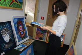 Pokaż zdjęcie: Zdjęcie korytarza biurowego, na krzesłach i podłodze poustawiane są prace plastyczne, kobieta trzyma w ręku obraz i go oglądaety i mężczyzna